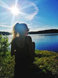 Rear view of young woman sitting by lake against blue sky during sunny day