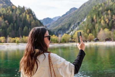 Side view of beautiful young woman using mobile phone, taking photos of beautiful lake