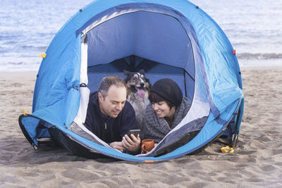 Man with woman using phone while relaxing by dog in tent at beach