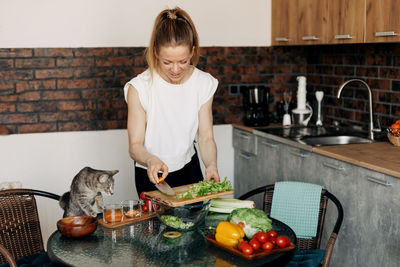 Young woman preparing vegetarian green salad at home and playing with cat