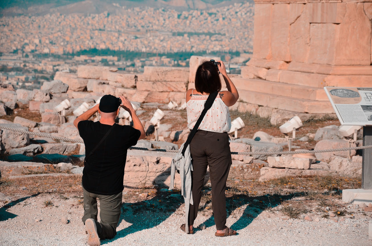 REAR VIEW OF WOMAN PHOTOGRAPHING WITH UMBRELLA STANDING ON COBBLESTONE