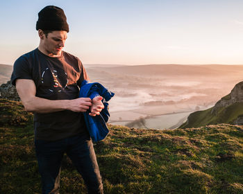 Man holding jacket while standing on mountain against sky during sunset