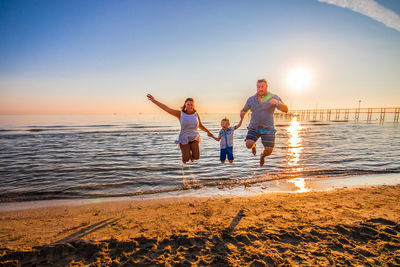 Parents holding son hands while jumping on shore at beach against sky during sunset