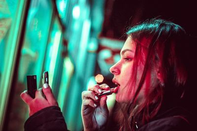 Side view of woman applying lipstick while looking in smart phone
