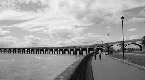 Rear view of people walking on road by sea against cloudy sky