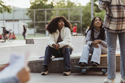 Teenage girls sitting on retaining wall looking at female friend in skateboard park