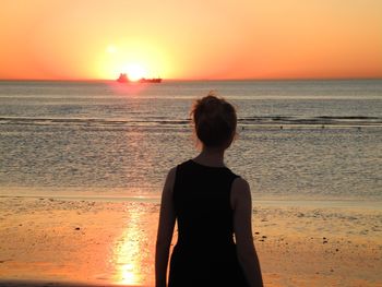 Rear view of woman standing at beach against orange sky