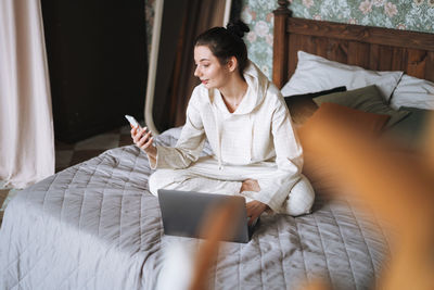 Beautiful smiling woman freelancer with dark long hair in casual using laptop sitting on bed at home