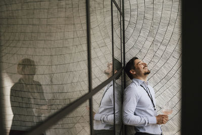 Cheerful businessman with head back discussing with female colleague while standing by glass wall