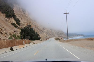 Empty road by mountains against sky during foggy weather seen through car windshield