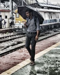 Young man carrying umbrella while walking on railroad platform during rainy season