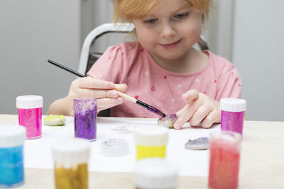 Little smiling child painting pebbles with colorful paints on the table.