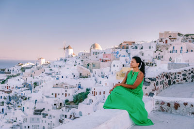 Woman sitting by buildings against clear sky