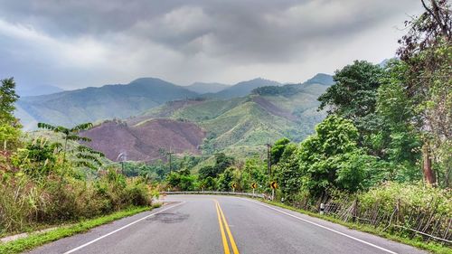 Empty road along trees and mountains against sky