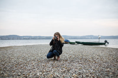 Woman photographing through camera while crouching at lakeshore against clear sky