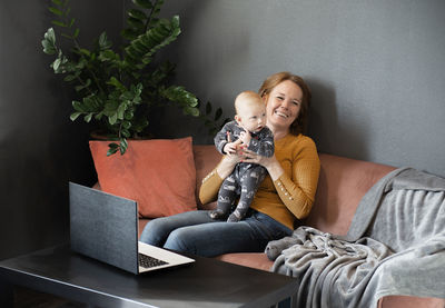 A happy grandmother plays with baby grandson on sofa at home living room,laptop on table
