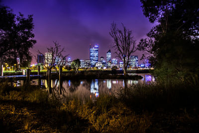 Illuminated city by lake against sky at night