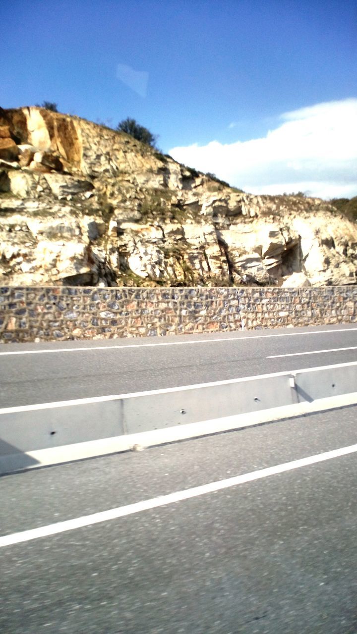 sky, blue, road, mountain, sunlight, road marking, rock - object, rock formation, day, transportation, no people, outdoors, asphalt, nature, built structure, cloud, the way forward, clear sky, shadow, street