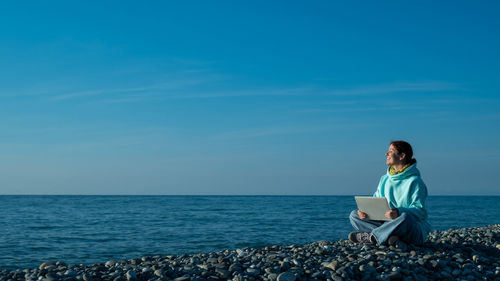 Caucasian woman working on a laptop on a pebbly seashore.