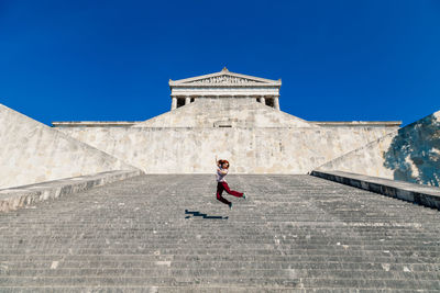 Low angle view of happy woman jumping on staircase against clear blue sky