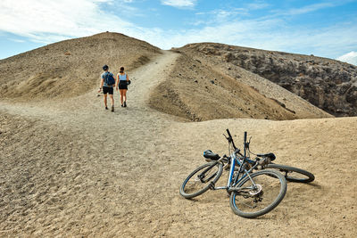 Unrecognizable man and woman walking away from bicycles on sandy path leading to dry hill on weekend day in fuerteventura, spain