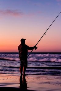 Silhouette man fishing on beach against sky at sunset