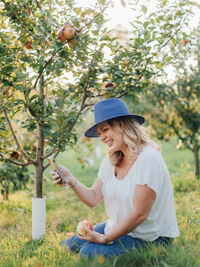 Smiling young woman holding hat while sitting on tree