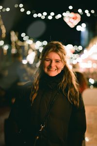 Portrait of young woman standing against illuminated string lights at night. 