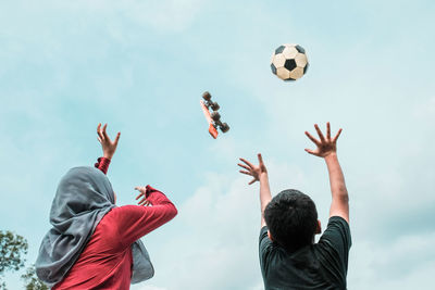 Rear view of siblings with arms raised against skateboard and soccer ball in mid-air