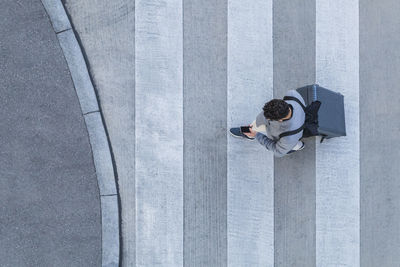 Businessman with baggage crossing the street while looking at cell phone, top view