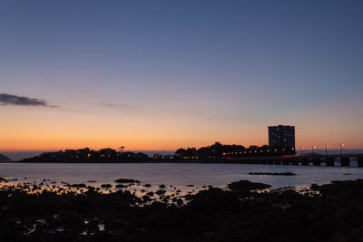Silhouette buildings by sea against sky during sunset