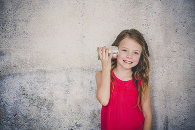 Cute smiling girl playing with tin can phone while standing against wall