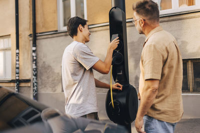 Rear view of father standing with son doing payment through smart phone at electric vehicle charging station