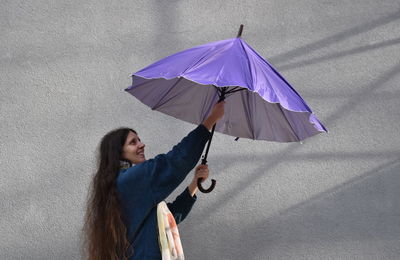 Side view of woman opening purple umbrella while standing against gray wall