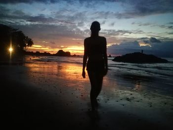 Rear view of silhouette woman standing on beach against sky during sunset