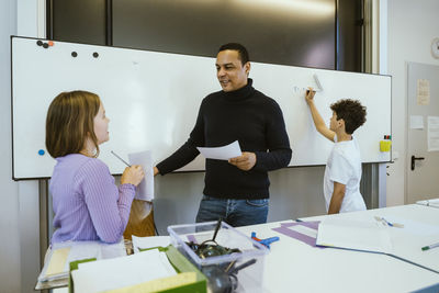 Male teacher assisting school students standing near whiteboard in classroom