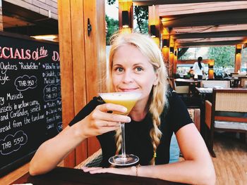 Portrait of smiling woman holding drink while sitting on table at cafe