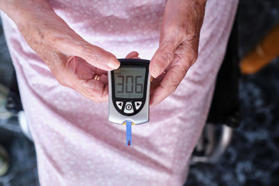 Top view of crop anonymous sick elderly female with special medical glucose meter in hands sitting in wheelchair in room