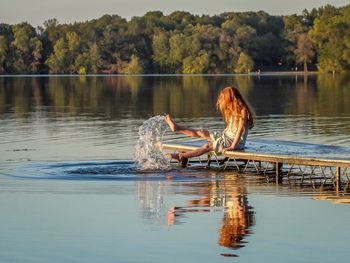 Teenager splashing lake water while sitting on jetty against trees