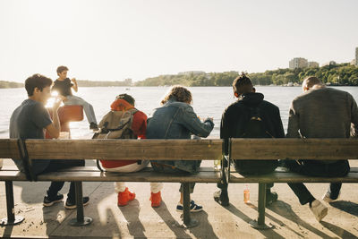 Rear view of people sitting on bench at shore against sky