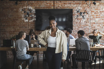 Female entrepreneur standing with arms crossed while colleagues working in creative office