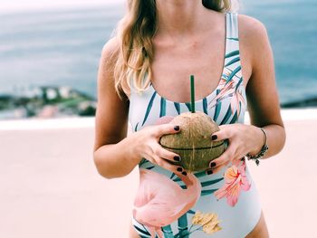 Midsection of woman in swimwear holding coconut at beach