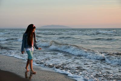 Girl walking at sea shore against sky during sunset