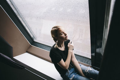 Young woman looking away while sitting on window