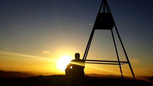 Man sitting by tripod against sky during sunset