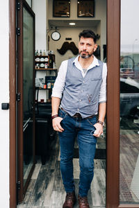 Adult bearded male beauty master with hands in pockets looking at camera at door of barbershop