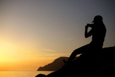 Silhouette woman photographing with mobile phone while sitting on rock against sky during sunset