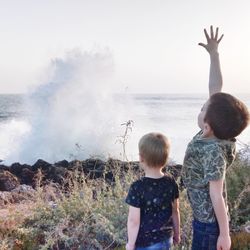 Rear view of boy with arms raised standing by brother near sea