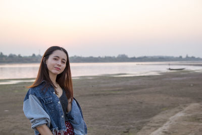 Portrait of young woman standing on beach against sky during sunset