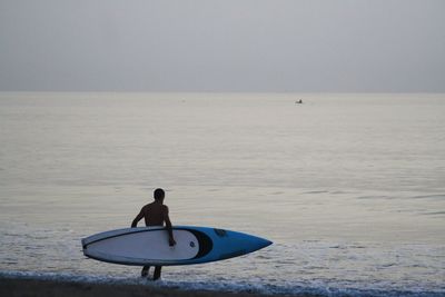 Rear view of man carrying surfboard on shore against sky